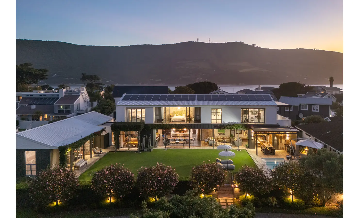 Perfect Hideaways aerial dusk view of an elegant luxury home with illuminated interiors, manicured garden, and surrounding residential rooftops against a mountain backdrop, The Pearl, Leisure Isle