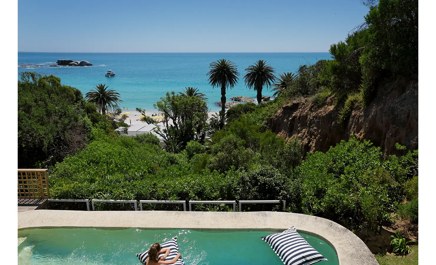 Perfect Hideaways a woman relaxing on striped float cushions in a curved pool overlooking lush greenery, tall palm trees, white sand beach, turquoise ocean and offshore boulders with a small boat anchored nearby, Clifton, Bungalow Les Lions