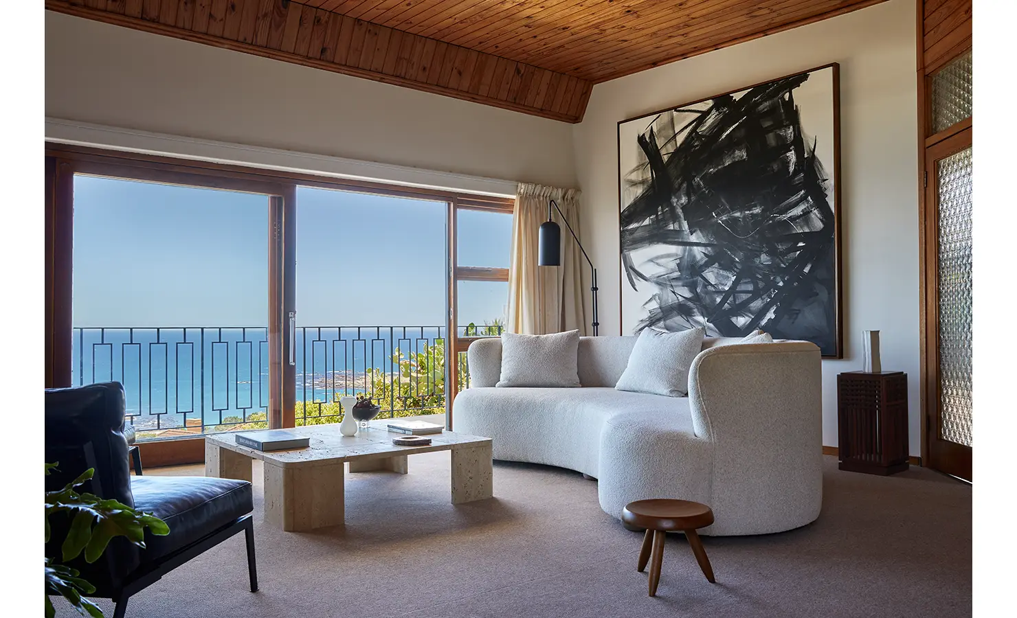 Perfect Hideaways a serene living room with a curved cream sofa, minimalist travertine coffee table, bold black abstract artwork, and expansive ocean views framed by wide glass doors beneath a warm wooden ceiling Camps Bay, The Liminal