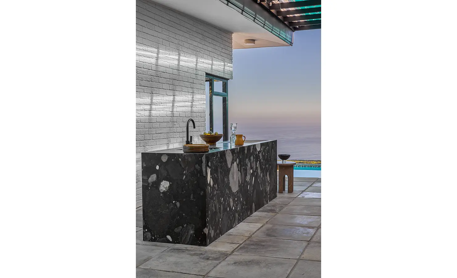 Perfect Hideaways an outdoor bar area featuring a dramatic black stone counter with large natural patterns, set against a white brick wall and overlooking a tranquil ocean horizon under a slatted pergola Camps Bay, The Liminal
