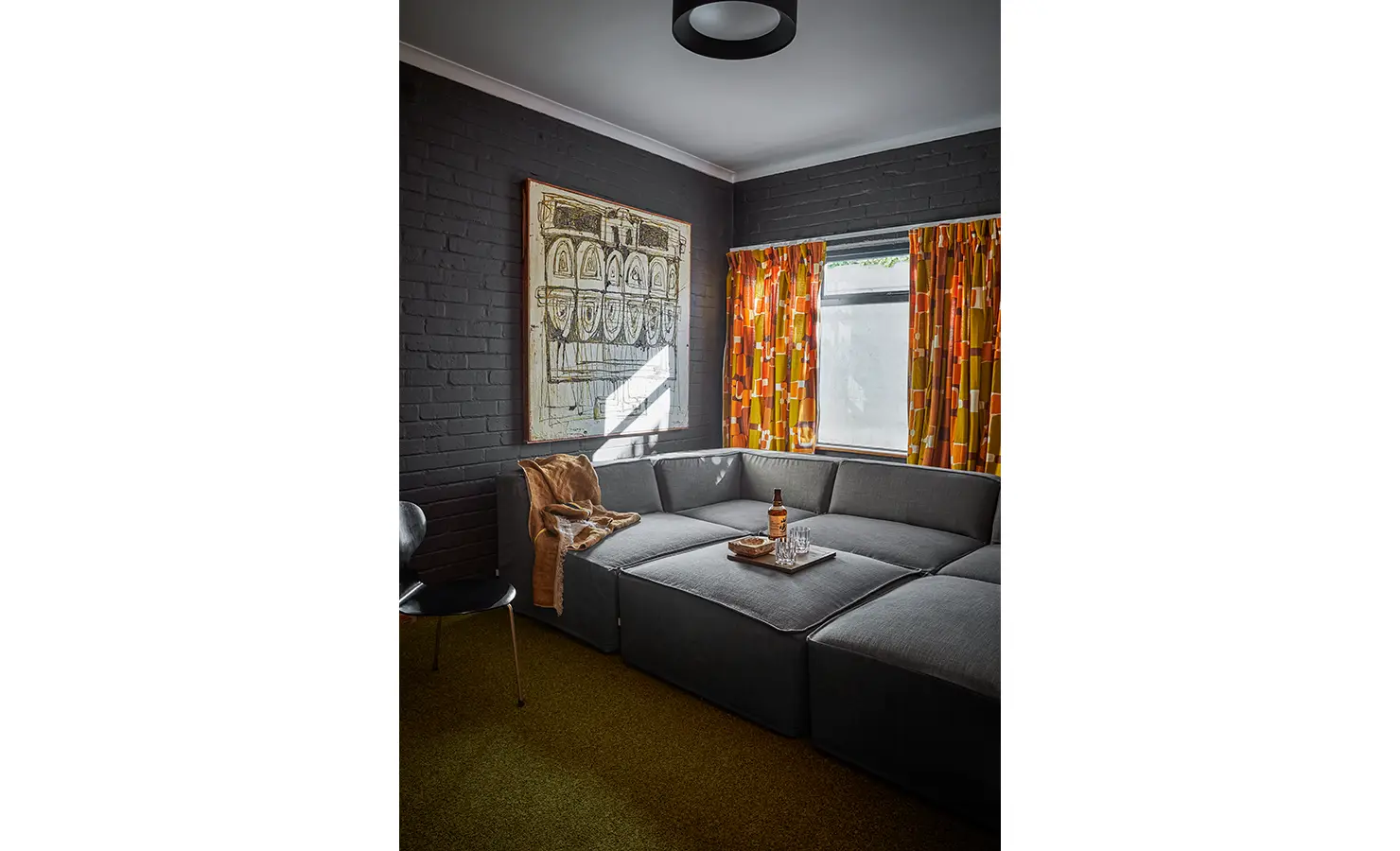 Perfect Hideaways a cozy TV lounge with dark brick walls, a modular grey sofa, retro patterned curtains, warm lighting, and a tray of drinks styled for relaxation Camps Bay, The Liminal