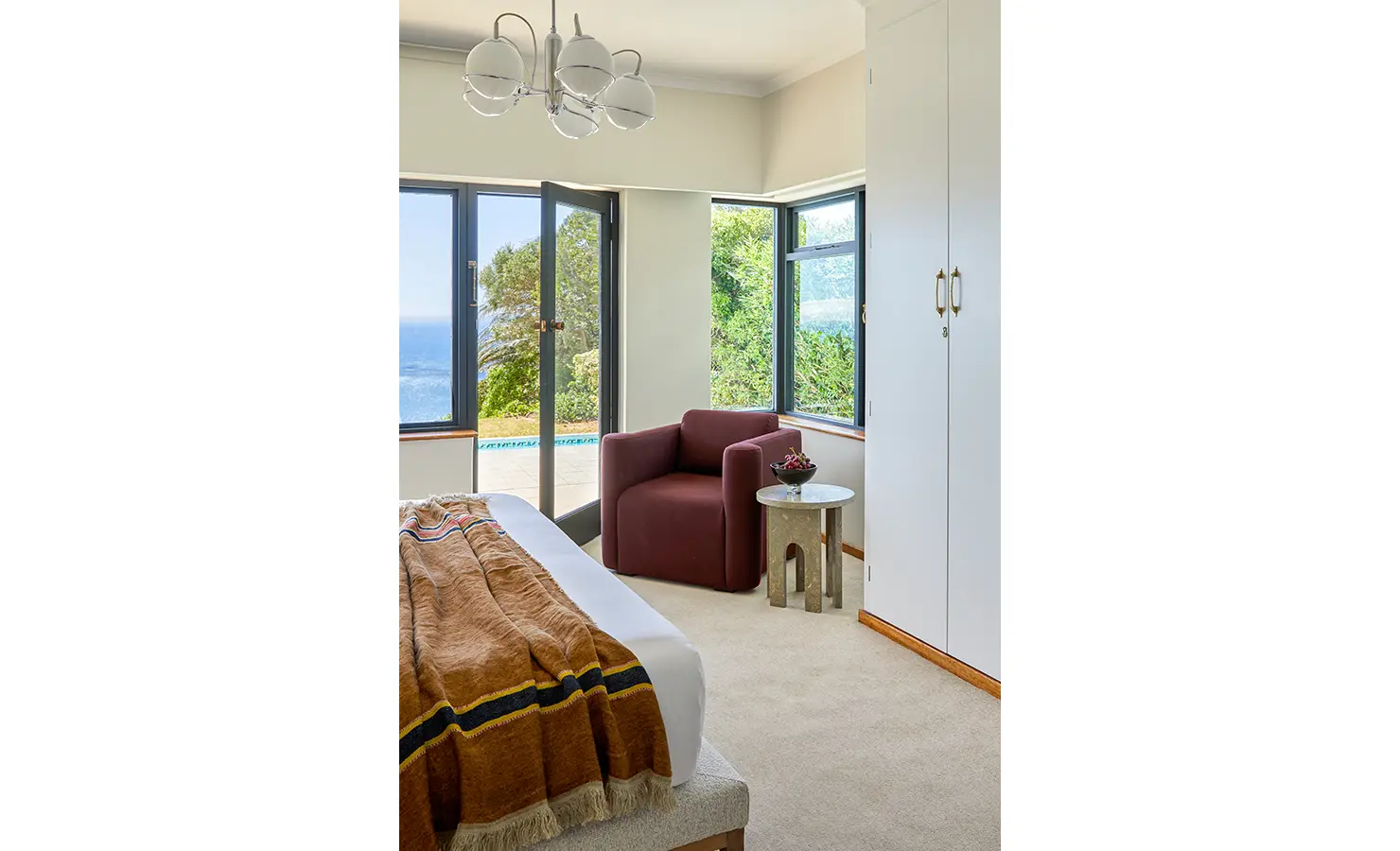 Perfect Hideaways a bright bedroom corner with a deep burgundy armchair beside a sculptural stone side table, surrounded by large windows framing lush greenery and a sparkling ocean view just outside the open glass door Camps Bay, The Liminal