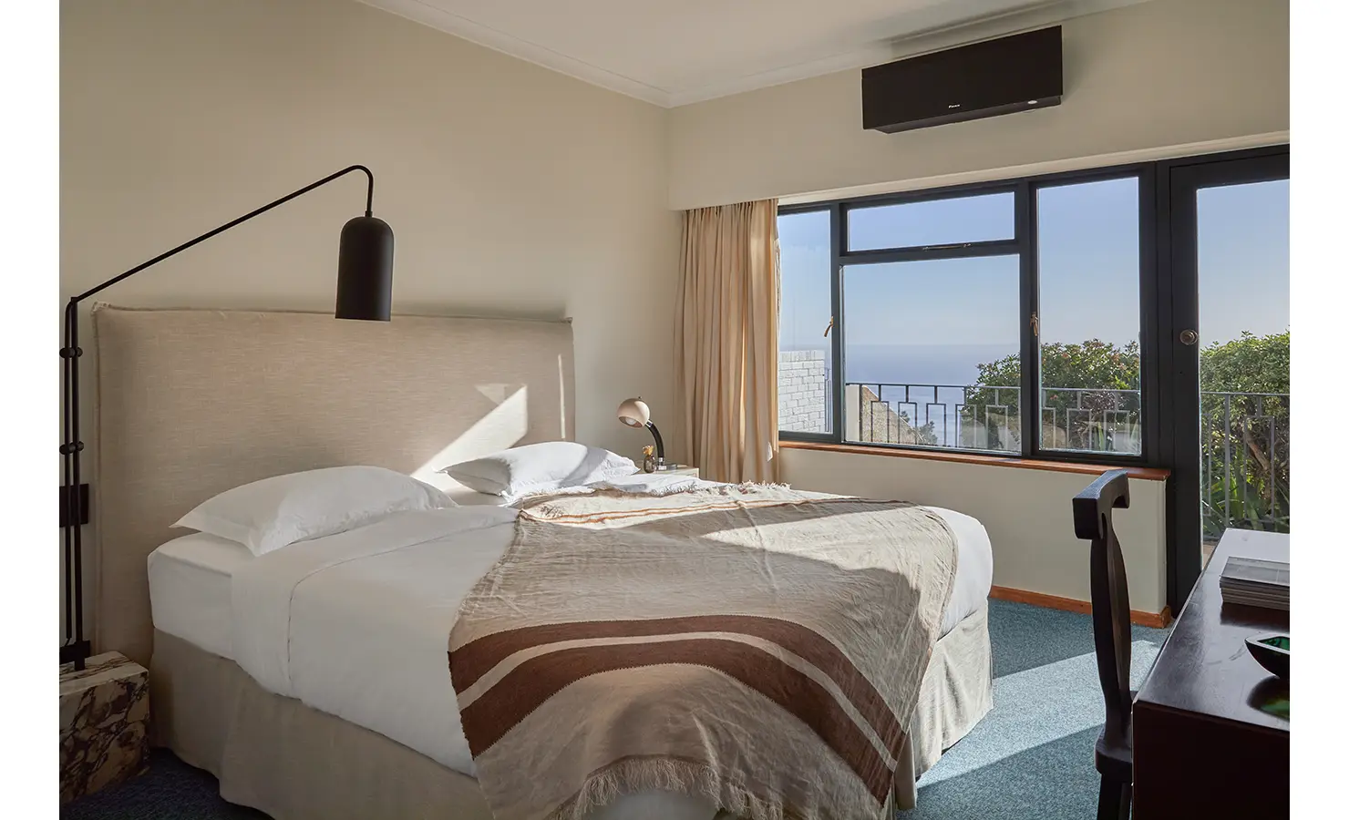 Perfect Hideaways a sunny bedroom with a tall upholstered headboard, neutral bedding, warm-toned throw blanket, and large windows overlooking greenery and the distant ocean Camps Bay, The Liminal