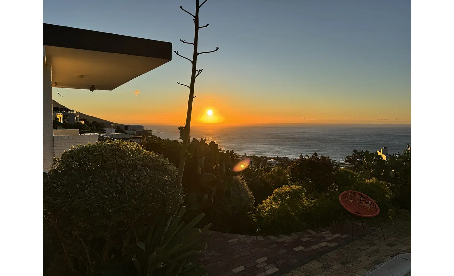 Perfect Hideaways a breathtaking sunset view over the Atlantic Ocean, casting warm golden light across lush garden foliage, distant rooftops, and a single red chair placed on a brick patio Camps Bay, The Liminal