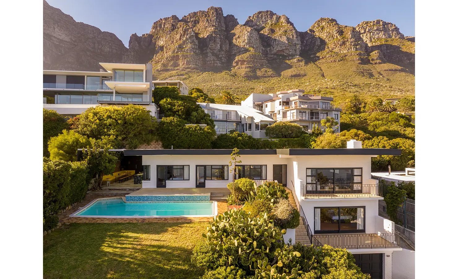 Perfect Hideaways an elevated exterior view of a modern white villa with clean mid-century lines, lush terraced greenery, a sparkling pool, and dramatic mountain cliffs rising directly behind the property Camps Bay, The Liminal
