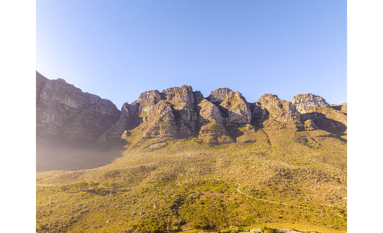 Perfect Hideaways a wide, sunlit landscape showing the rugged peaks and rocky contours of the Twelve Apostles mountain range under a clear blue sky, their warm tones glowing in the late afternoon light Camps Bay, The Liminal