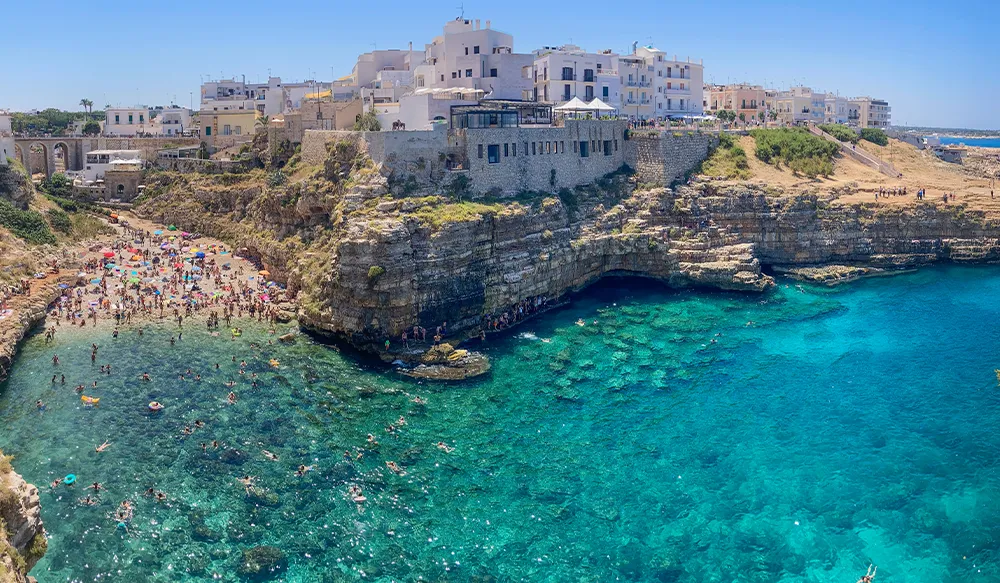 Perfect Hideaways blog aerial view of a dramatic cliffside cove in Polignano a Mare with whitewashed buildings perched above turquoise Adriatic waters and swimmers filling the rocky beach below, The heel of Italy