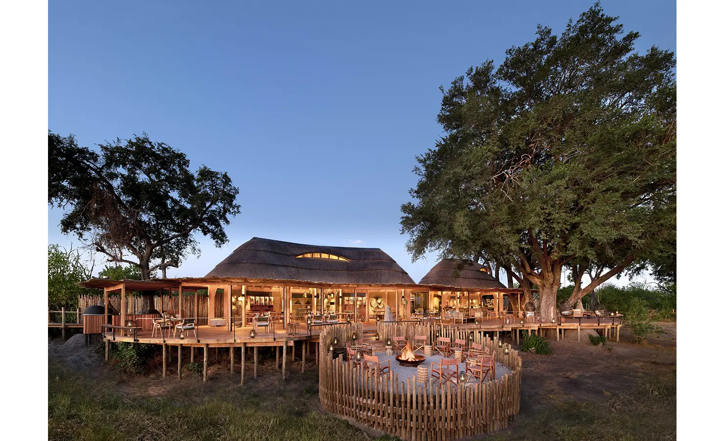 Perfect Hideaways elevated safari lodge with thatched roofs and open-air deck overlooking the bushveld with a central firepit at dusk, Tawana, Moremi Game reserve
