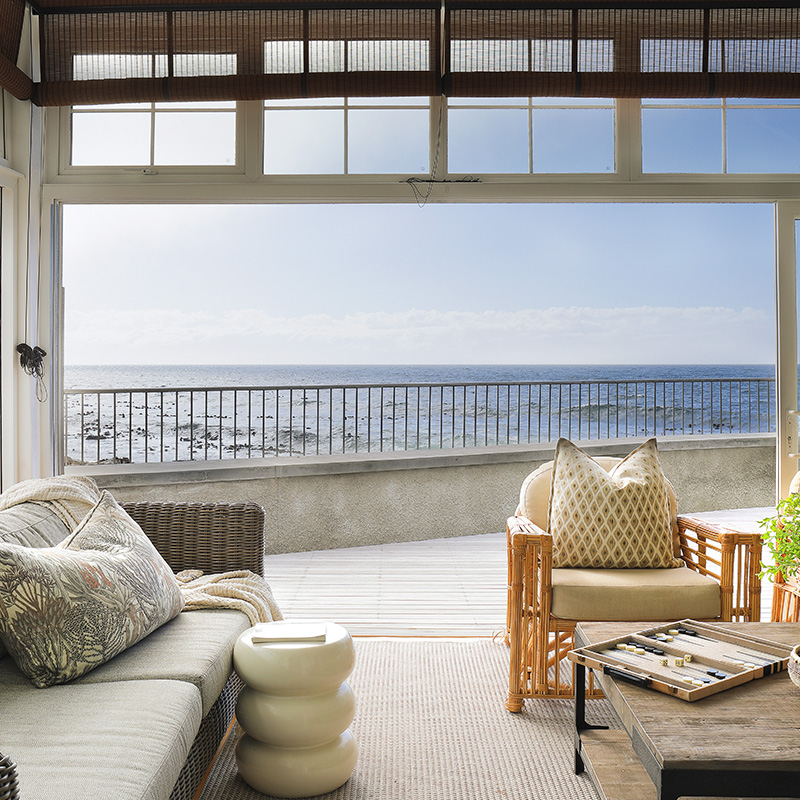 Perfect Hideaways coastal living room with woven sofa, patterned cushions and rattan chairs opening onto a sea-facing balcony with expansive ocean views, Maré House, Kalk Bay