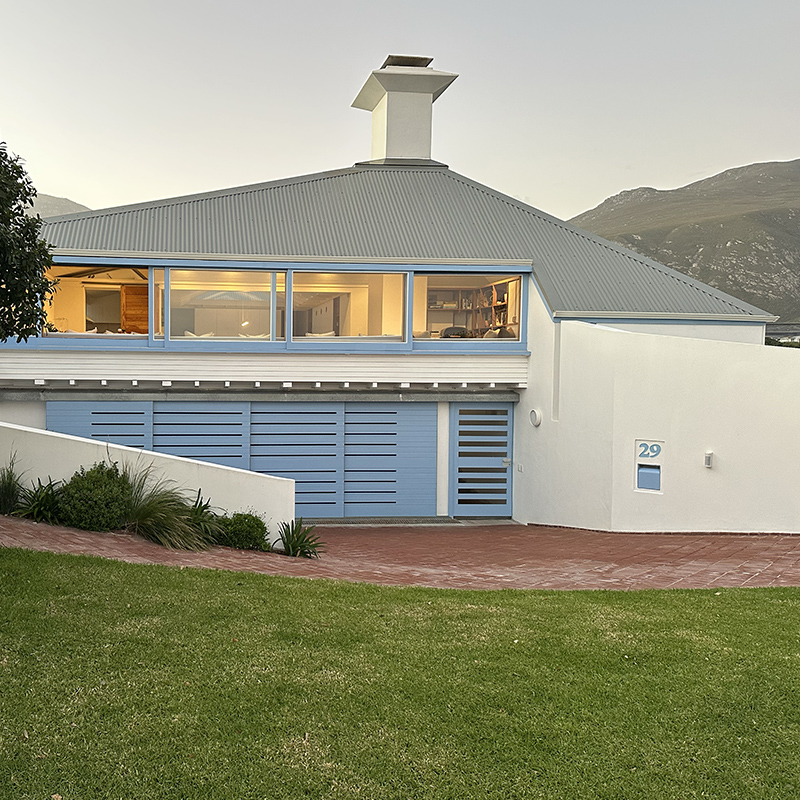 Perfect Hideaways contemporary coastal home with soft blue accents, large picture windows glowing warmly at dusk, landscaped garden and mountain backdrop, The Blue Nook, Hermanus