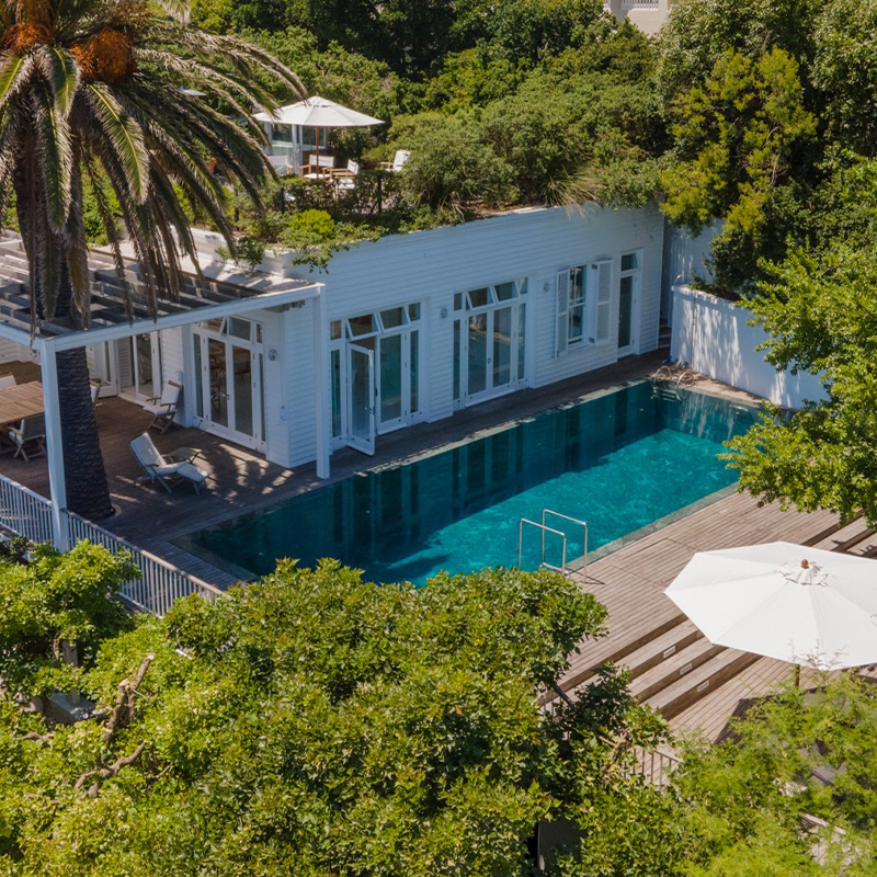 Perfect Hideaways contemporary white villa with expansive wooden deck, long swimming pool, and shaded outdoor dining beneath a large palm tree surrounded by lush greenery, Villa Oasis, Bantry Bay