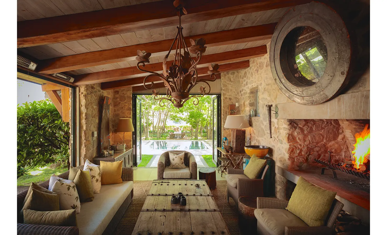Perfect Hideaways rustic indoor-outdoor lounge with stone fireplace, wooden beams, chandelier and comfortable seating opening onto garden and swimming pool, The Manor, Franschhoek