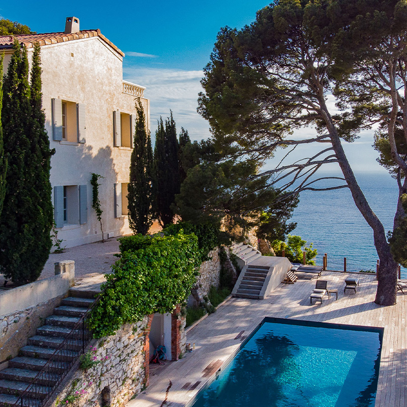 Perfect Hideaways: A serene Mediterranean villa with pale stone walls, shuttered windows, and tall cypress trees lining a terrace that overlooks a wooden deck with a sparkling pool, all perched above the deep blue sea framed by mature pines — Cassis de Luxe, Cassis