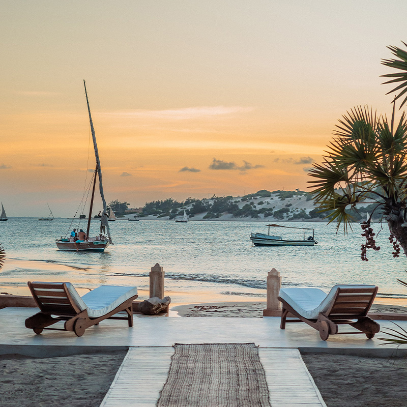 Perfect Hideaways beachfront terrace with two wooden loungers overlooking calm ocean waters at sunset, traditional dhow boats sailing past and palm trees framing the view, Syngue Sabour, Ras Kitau
