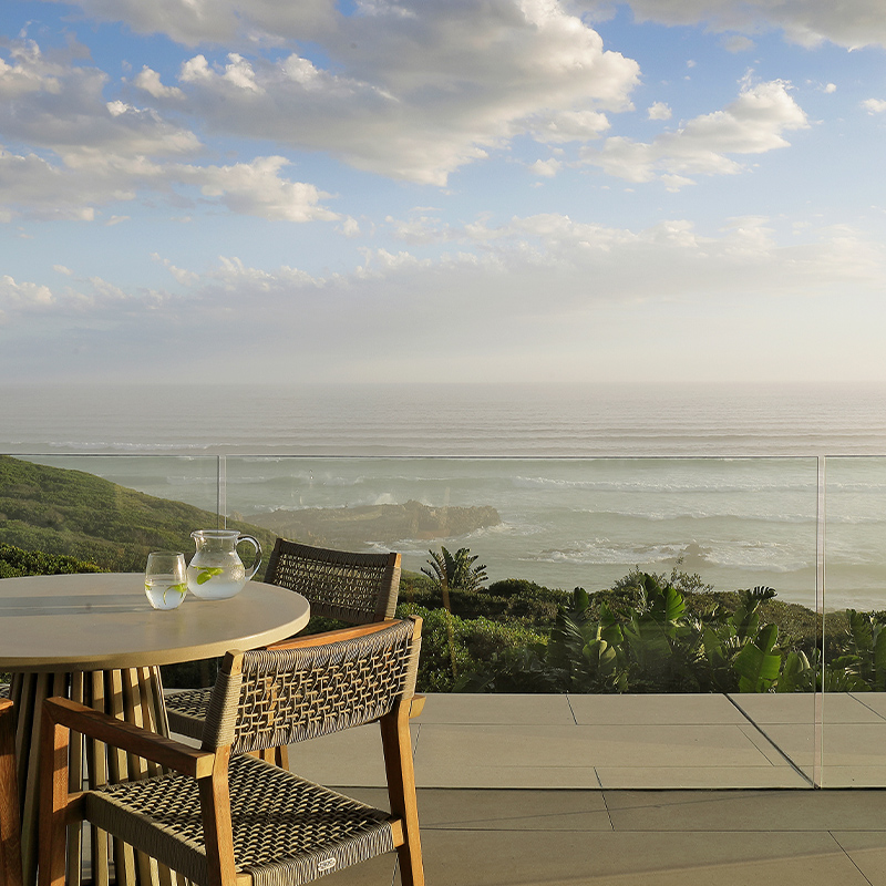 Perfect Hideaways ocean-view terrace with round dining table and woven chairs overlooking lush coastal greenery and expansive sea horizon at sunset, The 147, Hermanus