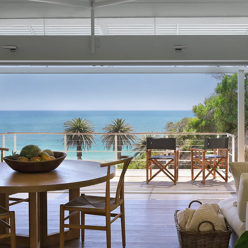 Perfect Hideaways: View from inside of the open plan lounge dining room with round wooden table and chairs looking out double stacked doors onto balcony and Clifton Beach sea views at Bungalow Les Lions Clifton.