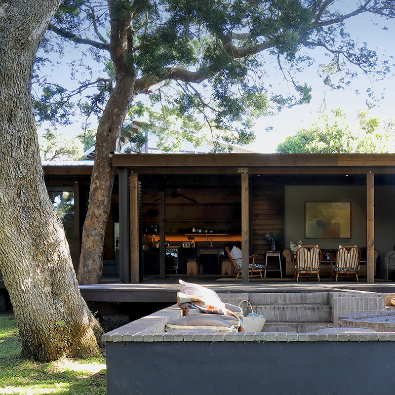 Perfect Hideaways contemporary timber retreat with outdoor fire pit, shaded deck and open-plan living space beneath mature trees at Lagoon Retreat Bosbok, Brenton-on-Lake